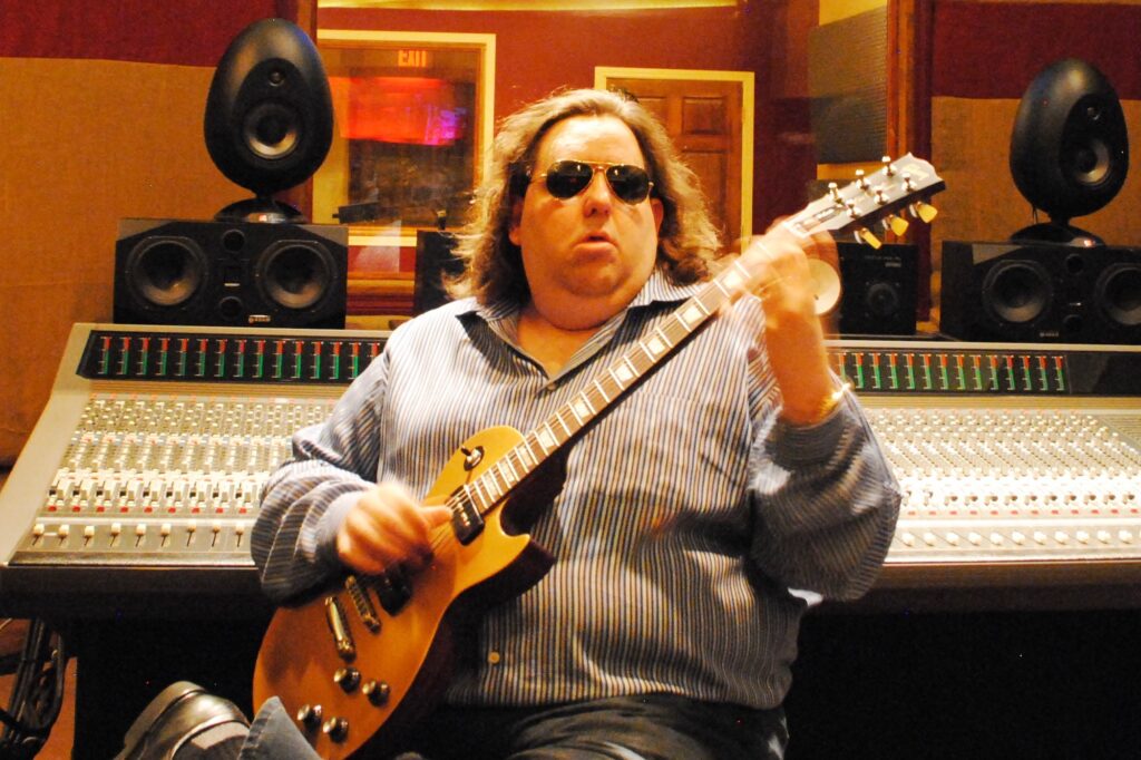 Joey Stuckey seated with a gold-top Les Paul electric guitar in front of the mixing desk at Shadow Sound Studio.